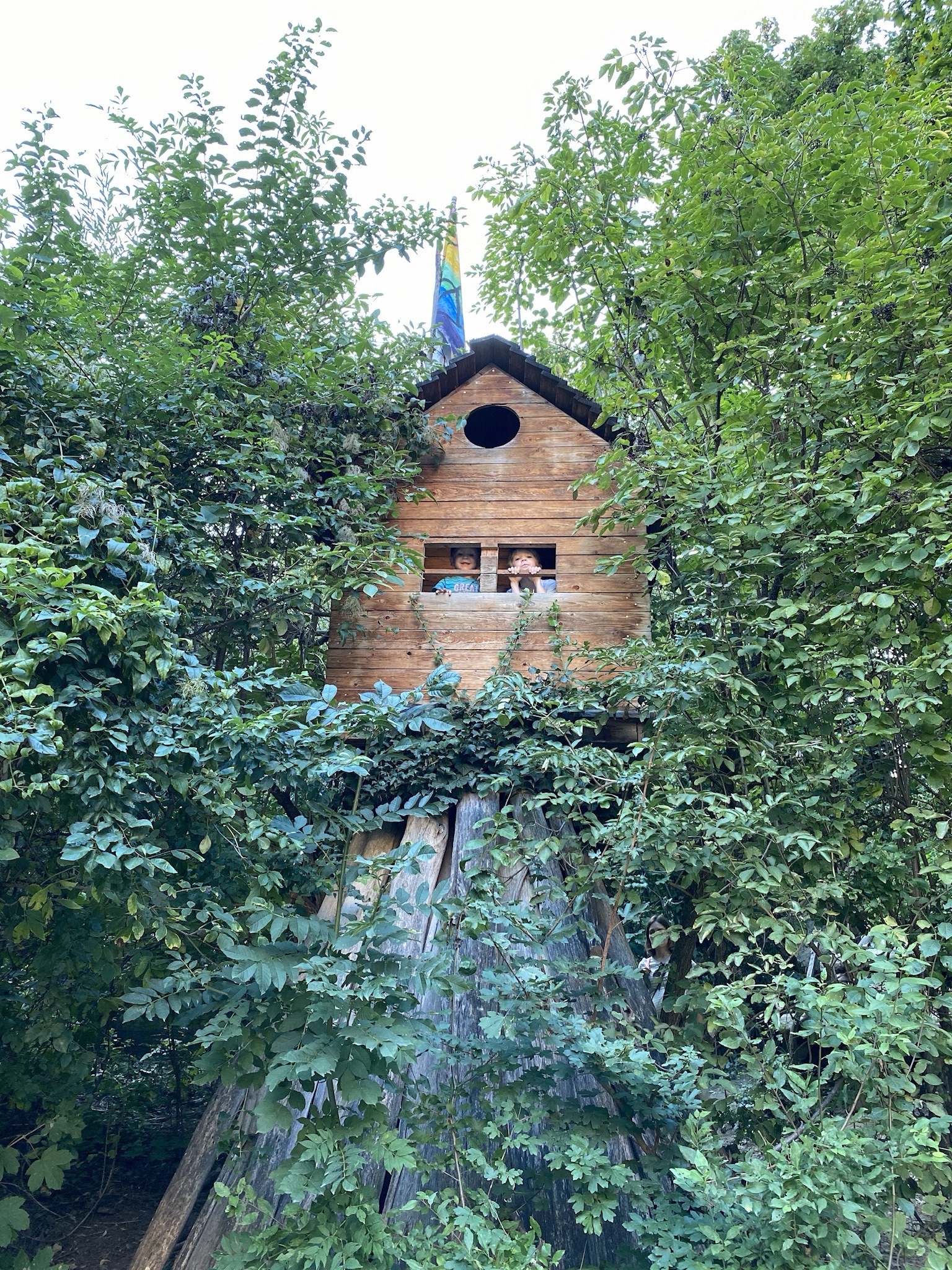 Two of my kids in the wooden playhouse surrounded by dense greenery at the Robinson playground